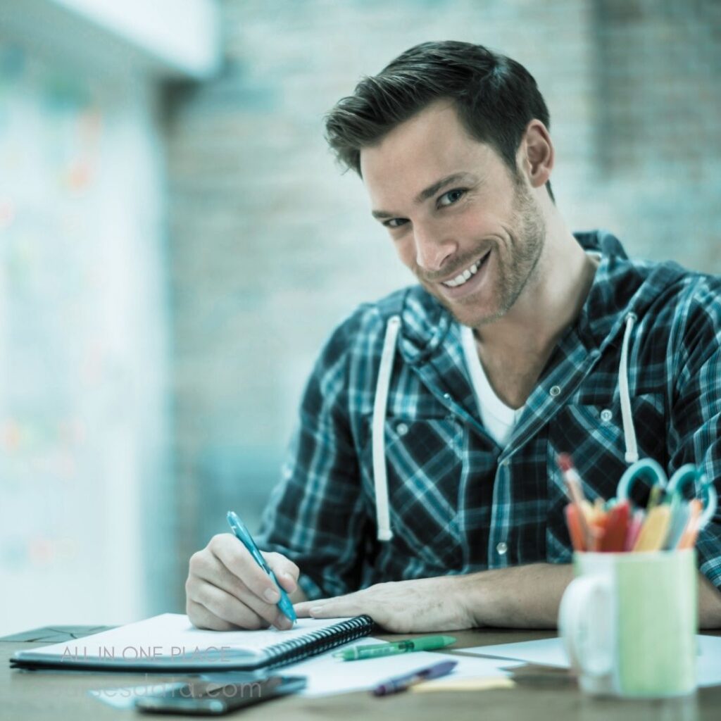 Smiling man at desk. Holding blue pen. Writing on notebook. Colorful stationery nearby. Casual plaid shirt.