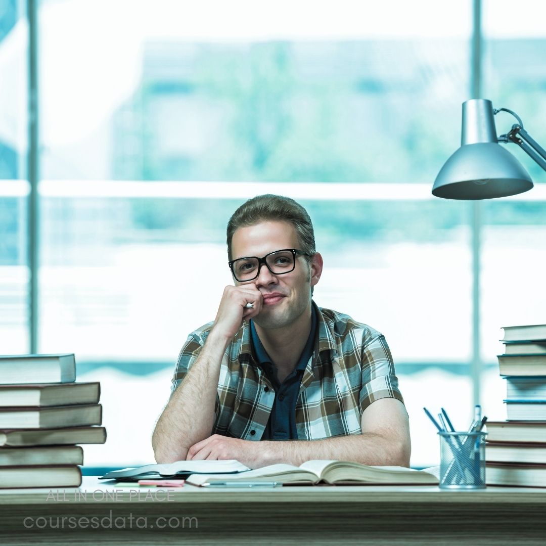 Man smiling at desk. Wearing glasses and plaid shirt. Surrounded by stacked books. Bright window in background. Desk lamp illuminating workspace.