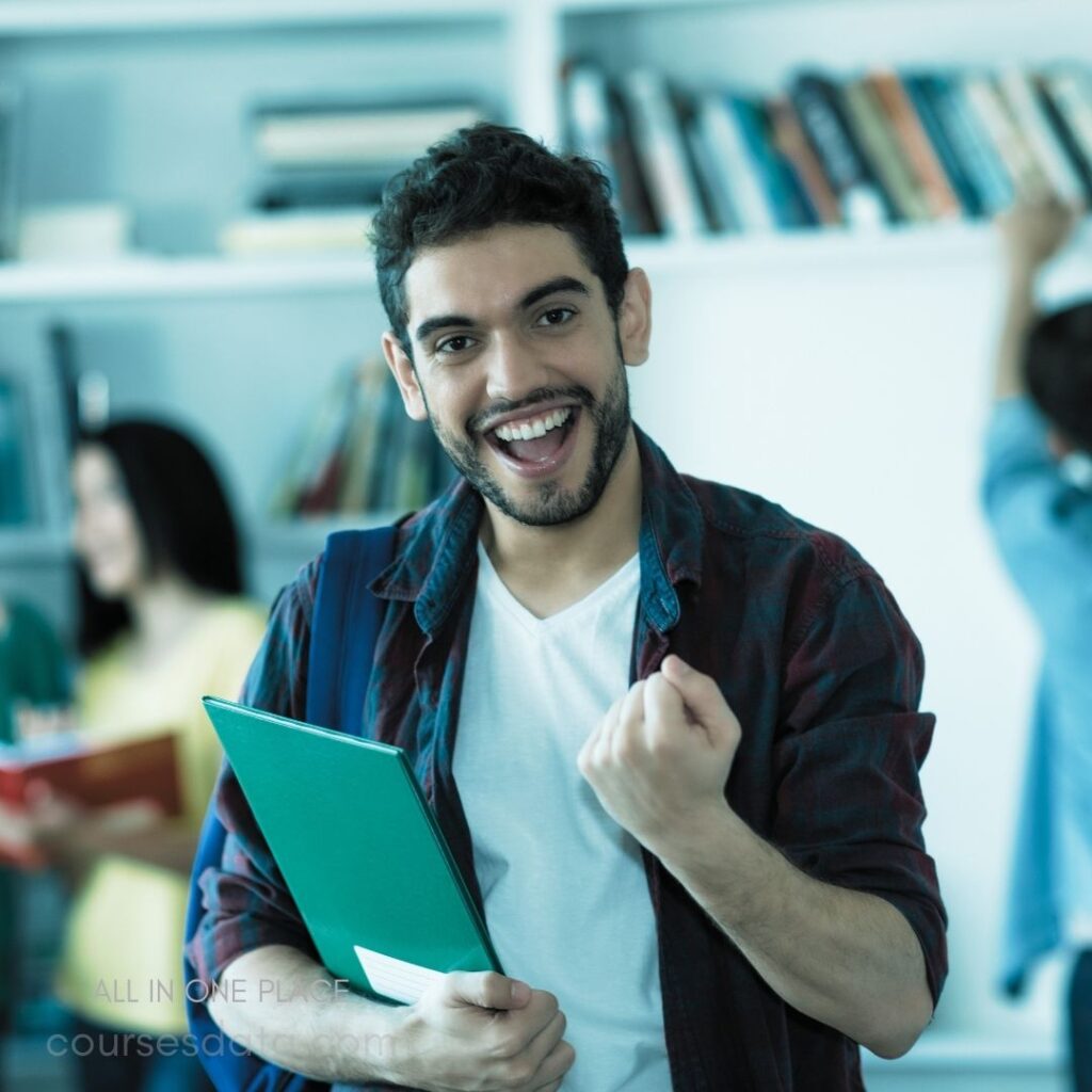 Smiling man with green folder. Joyful expression, fist raised. Bookshelves filled with books. People studying in background. Casual clothing, relaxed atmosphere.