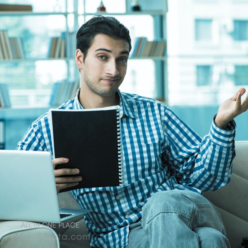 Man in plaid shirt. Holding a notebook. Sitting on a couch. Laptop on the table. Casual office environment.