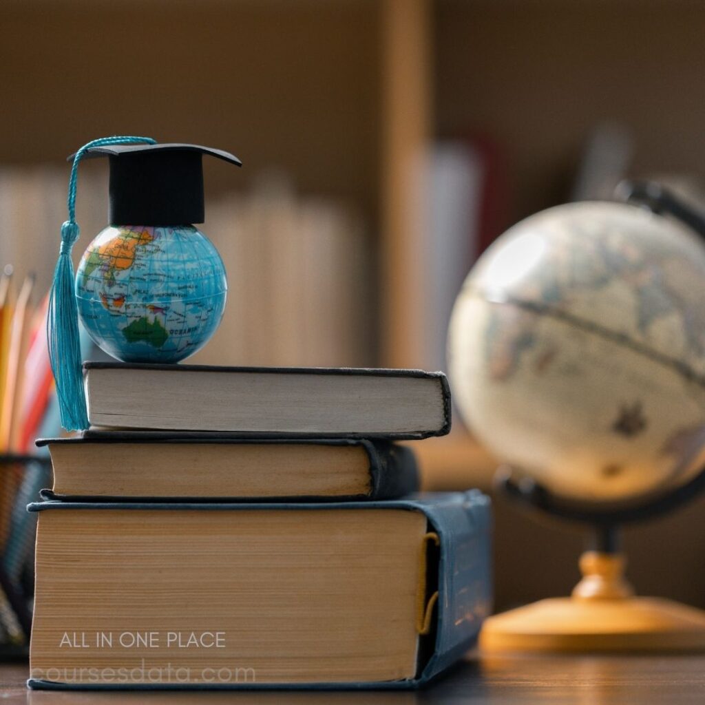 Mini globe with graduation cap. Stacked books beneath globe. Large globe in background. Organized study space setting. Colorful pencils in holder.