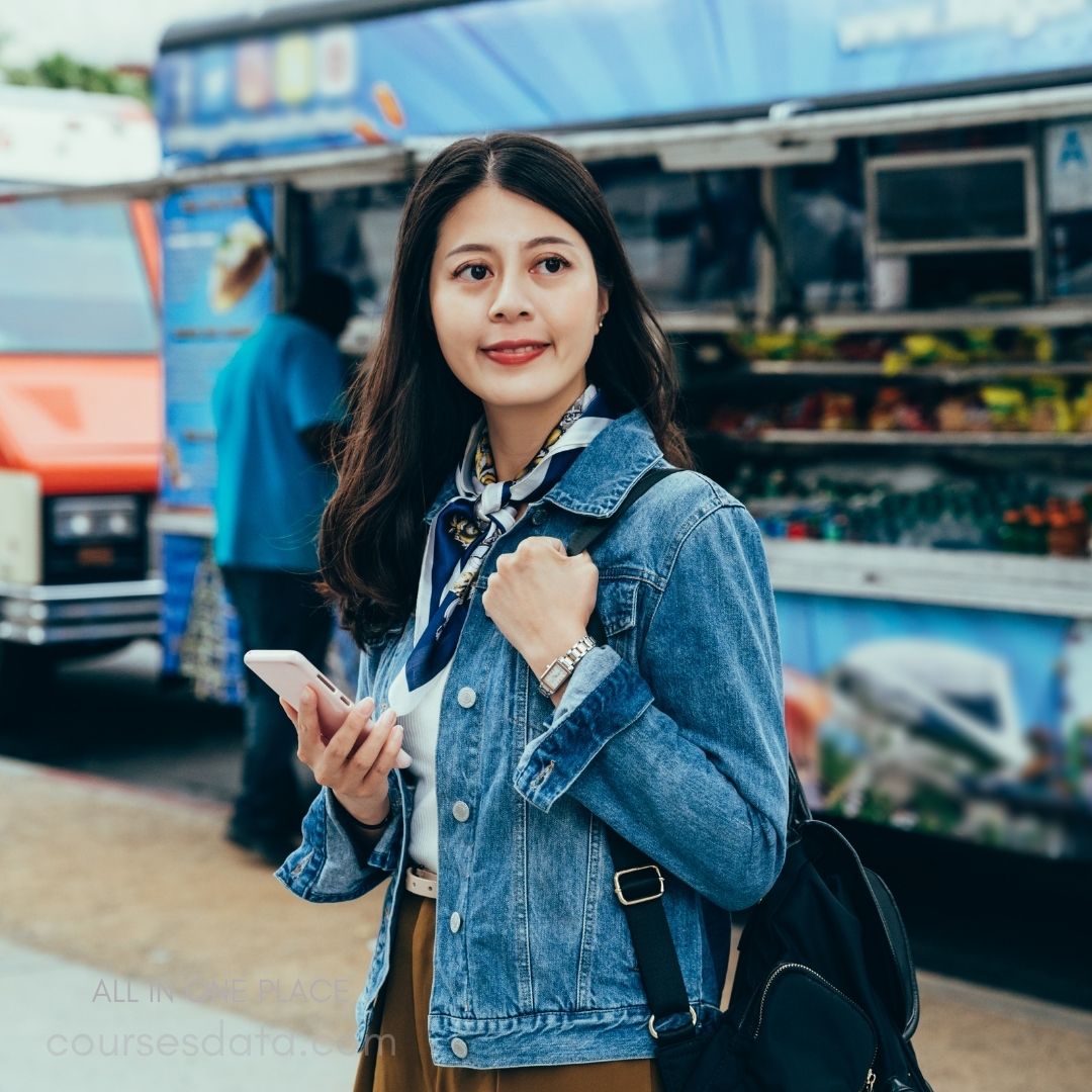 Young woman in denim jacket. Holding smartphone in hand. Wearing scarf and watch. Food truck background visible. Casual urban setting.