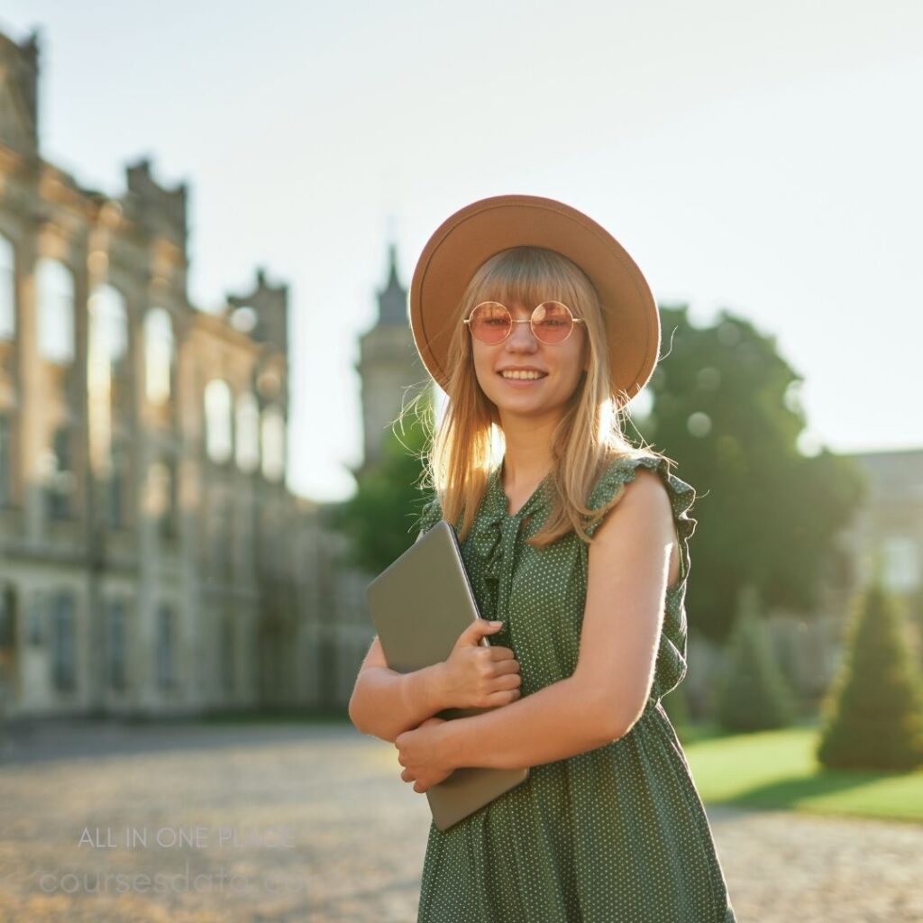 Smiling girl in summer dress. Wearing hat and sunglasses. Holding a laptop close. Historic building in background. Sunlit outdoor scene.