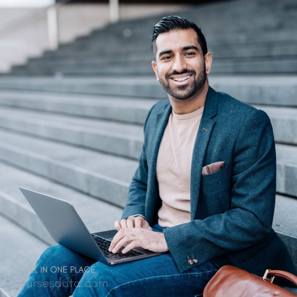 Smiling man on steps, laptop. Wearing a blazer and sweater. Brown leather bag beside him. Natural lighting, urban setting. Casual and professional vibe.