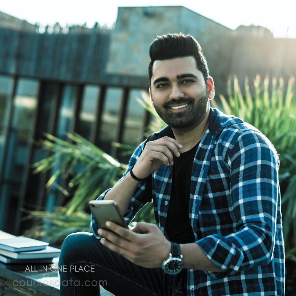 Smiling man holding smartphone. Wearing blue plaid shirt. Sitting outdoors with greenery. Sunlit background with reflections. Casual, relaxed atmosphere.