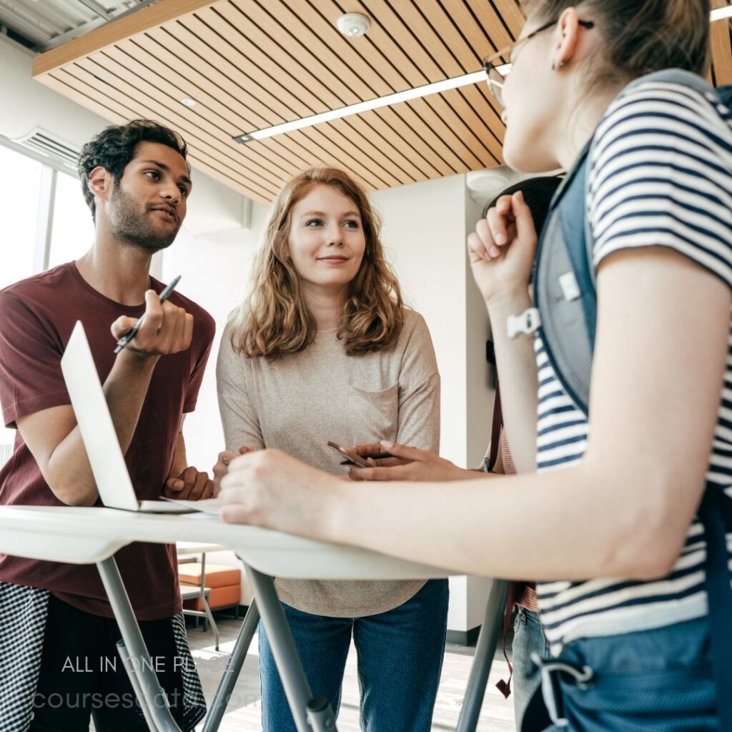 Group discussing around table. Young adults engaged in conversation. Laptop and pens on table. Casual attire in modern setting.