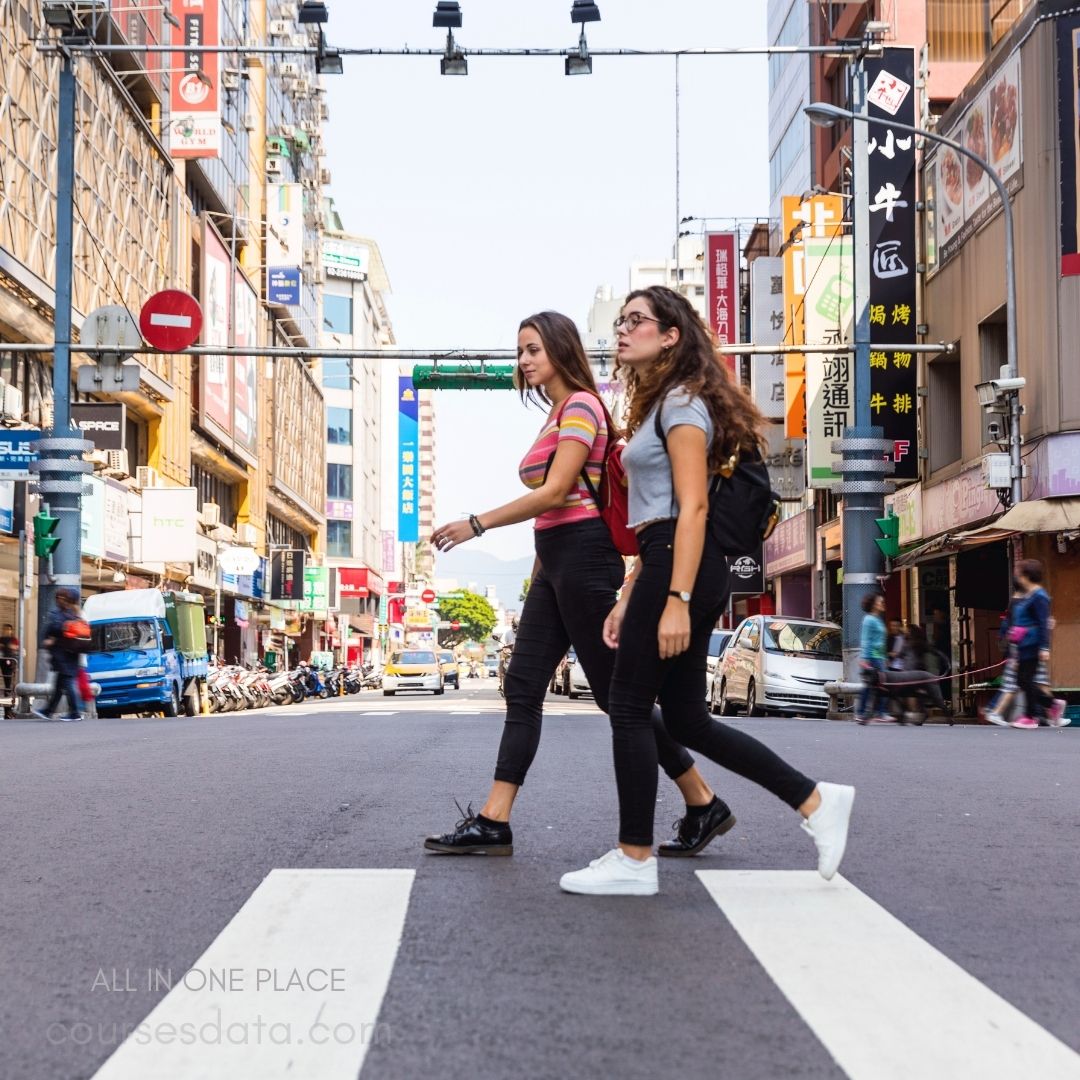 Two women crossing street. Colorful shops line street. Traffic lights visible overhead. Clear blue sky backdrop. Casual fashion, urban setting.