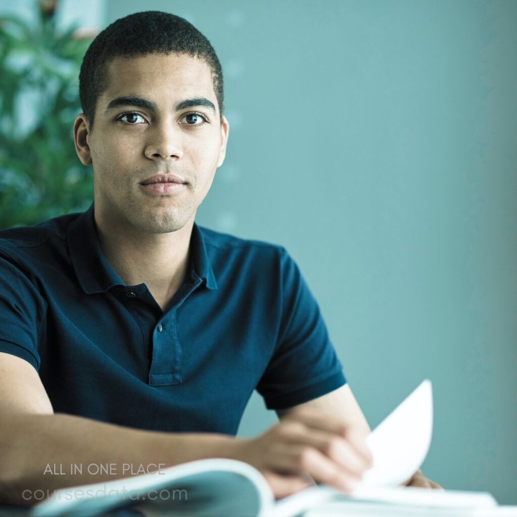 Young man, serious expression. Dark polo shirt. Sitting at a table. Open book in hands. Soft background with greenery.