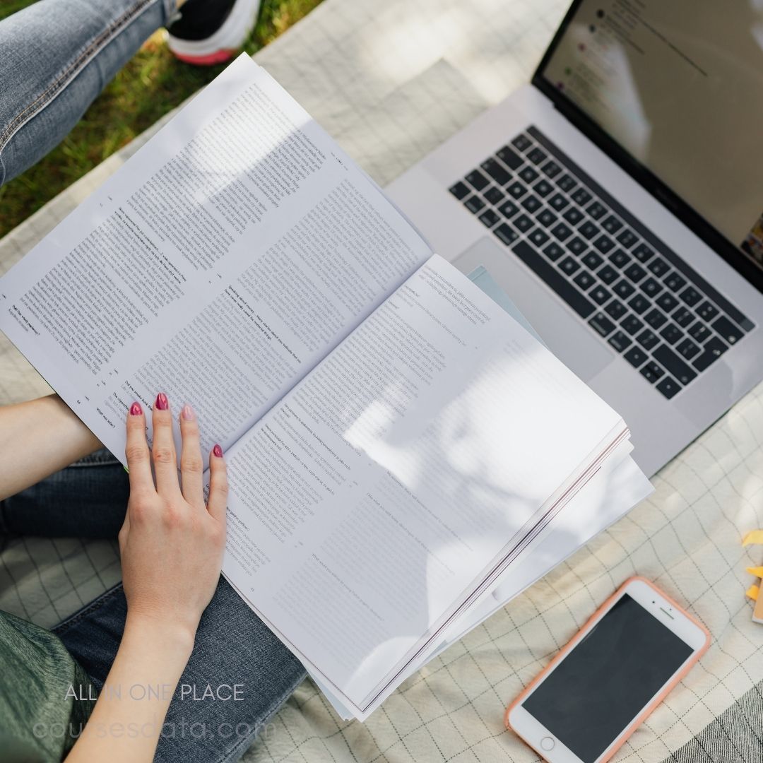 Reading a book outdoors, laptop nearby.