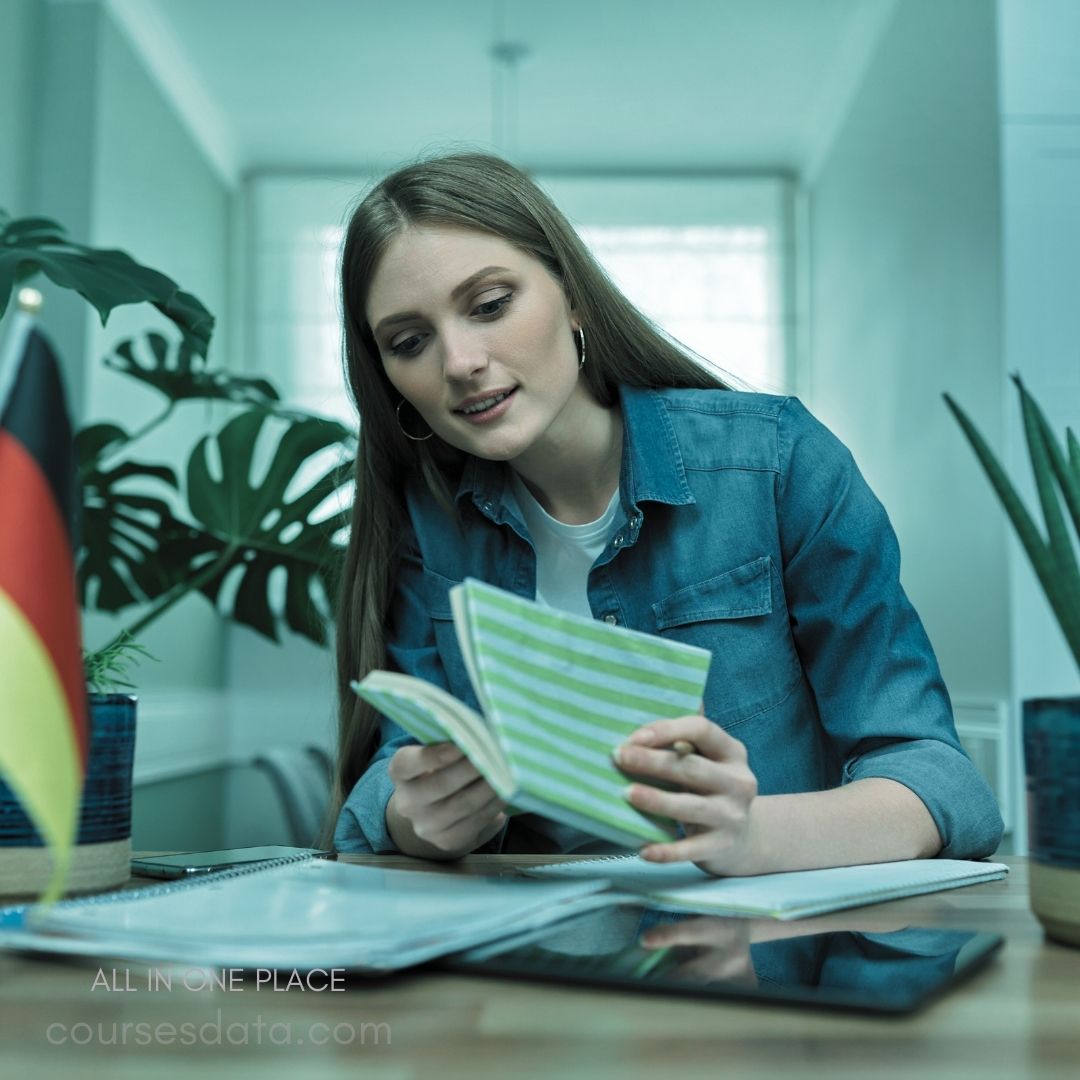 Woman reading from notebook. Green striped pages visible. German flag in background. Potted plant next to her. Study materials on table.