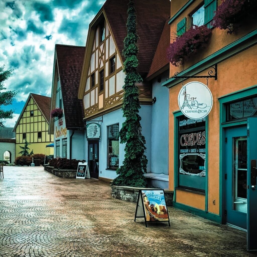 Quaint European-style shops, colorful facades. Cobblestone walkway, overcast sky. Green ivy climbing walls. Signs for crepes and café.