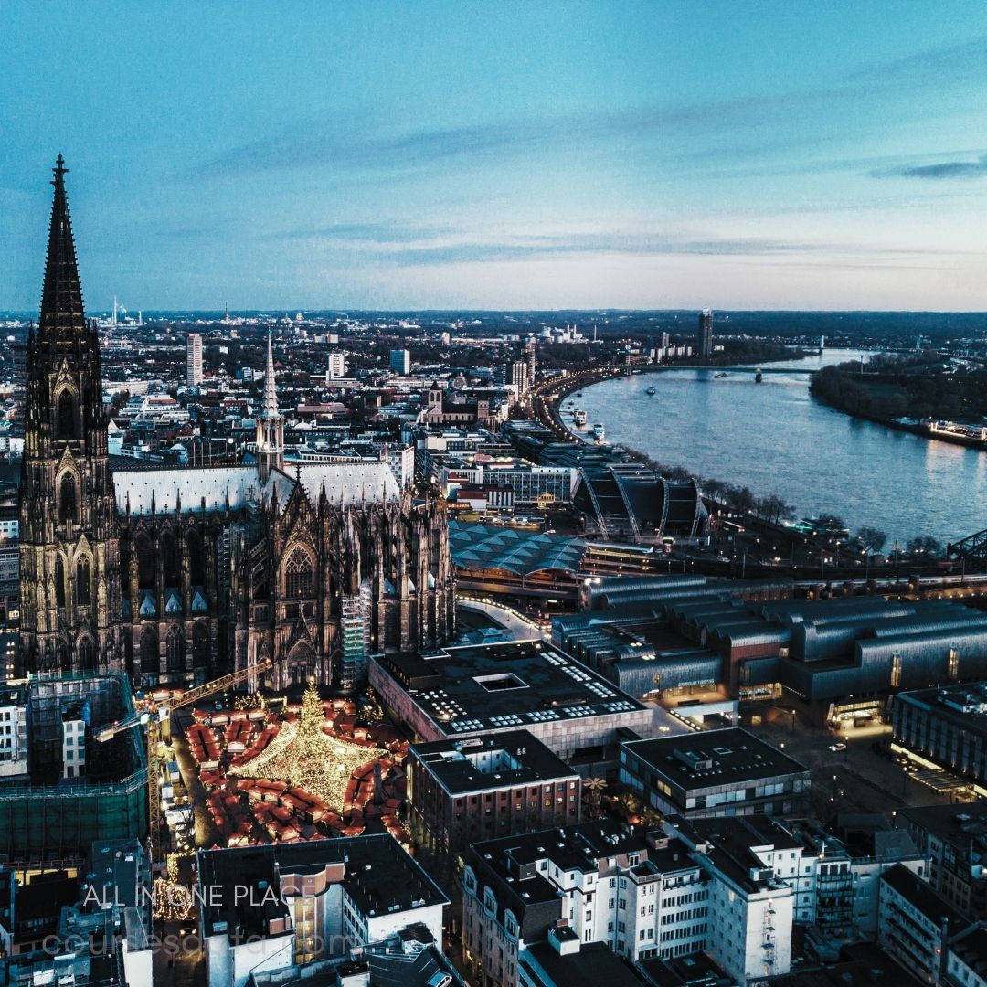Cologne Cathedral at dusk. Festive market illuminated below. Rhine river curving nearby. Cityscape with modern buildings. Twilight sky with gentle clouds.