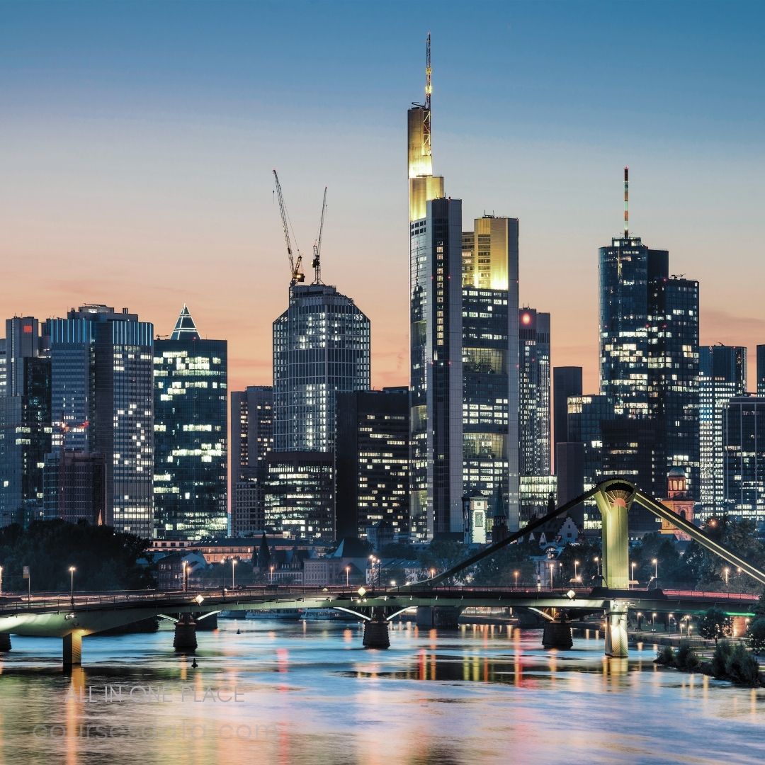 Frankfurt skyline at sunset. Illuminated skyscrapers and cranes. Bridge over reflective river. Clear sky with soft hues. City lights beginning to twinkle.