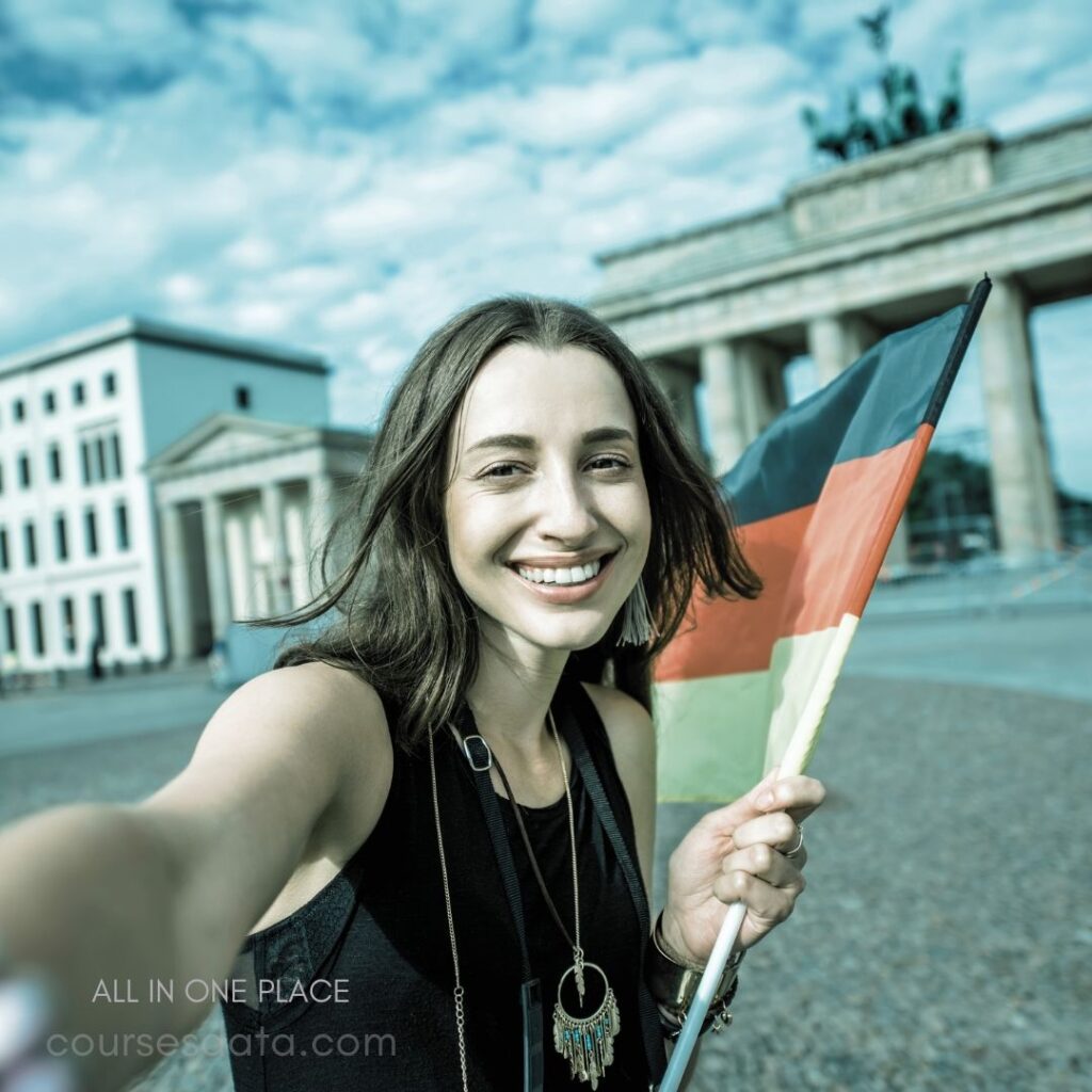 Smiling woman holding German flag. Berlin's Brandenburg Gate backdrop. Bright sky overhead, casual attire. Jewelry and selfie pose noticeable. Vibrant atmosphere and joyful expression.