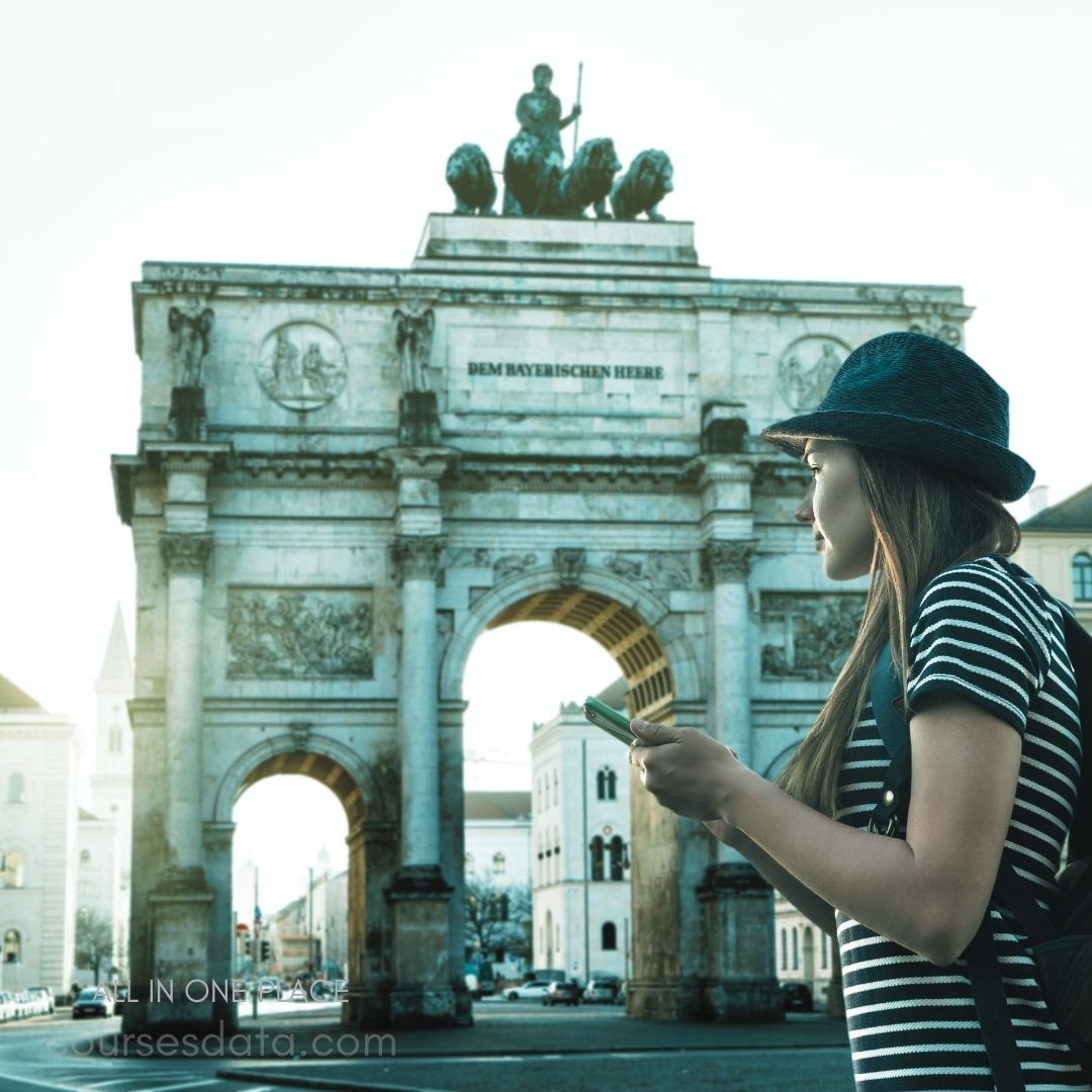 Woman by historical arch. Statue on top. Striped shirt and hat. Holding a smartphone. Morning light ambiance.