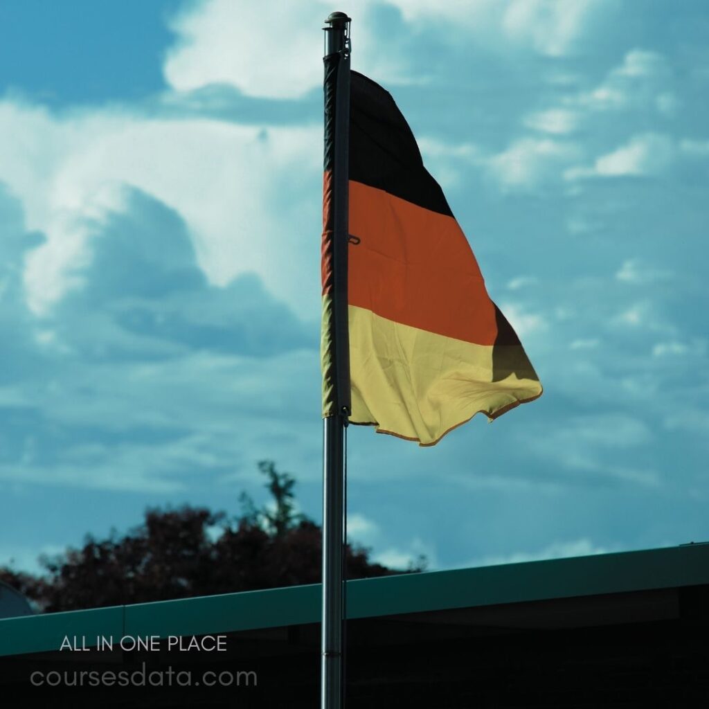 German flag waving proudly. Blue sky backdrop with clouds. Near a building structure. Flagpole anchored securely. Colors: black, red, gold.