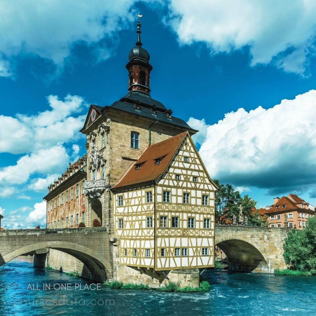 Historic building by river. Half-timbered architecture. Bridge over clear water. Bright blue sky, white clouds. Lush greenery nearby.