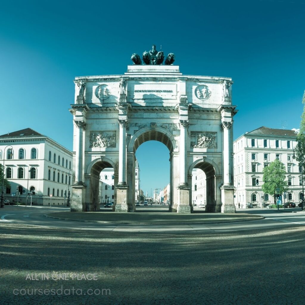 Triumphal arch in city center. Four pillars support structure. Statues atop arch. Clear blue sky backdrop. Surrounding buildings visible.