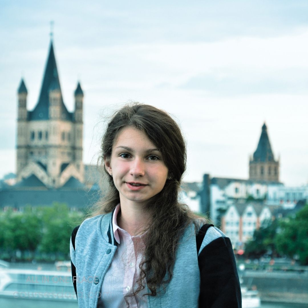 Young girl in a city. Historic towers in background. Overcast sky above. Casual attire, friendly expression. Greenery along waterfront.