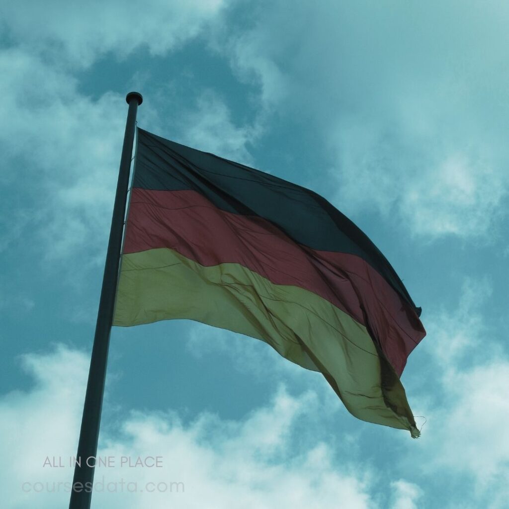 German flag waving gently. Cloudy blue sky backdrop. Black, red, and yellow stripes. Flagpole standing prominently. Symbol of national identity.