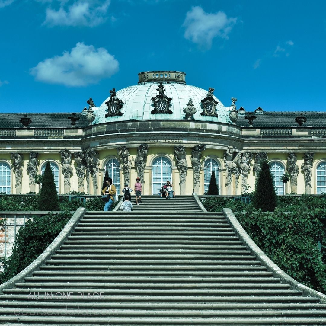 Historic building with dome. Stairs leading to entrance. People relaxing on steps. Greenery surrounding the structure. Clear blue sky backdrop.