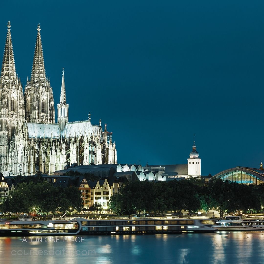 Cologne Cathedral at night. Illuminated gothic architecture. River reflections and cityscape. Historic buildings along waterfront. Peaceful evening atmosphere.