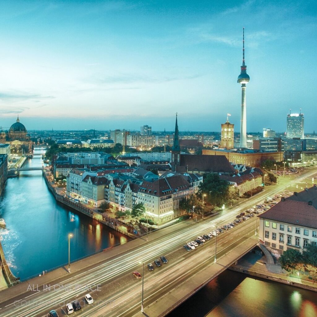 Berlin cityscape at twilight. TV tower in the background. Historic buildings along river. Street lights illuminate roads. Calm water reflecting skyline.