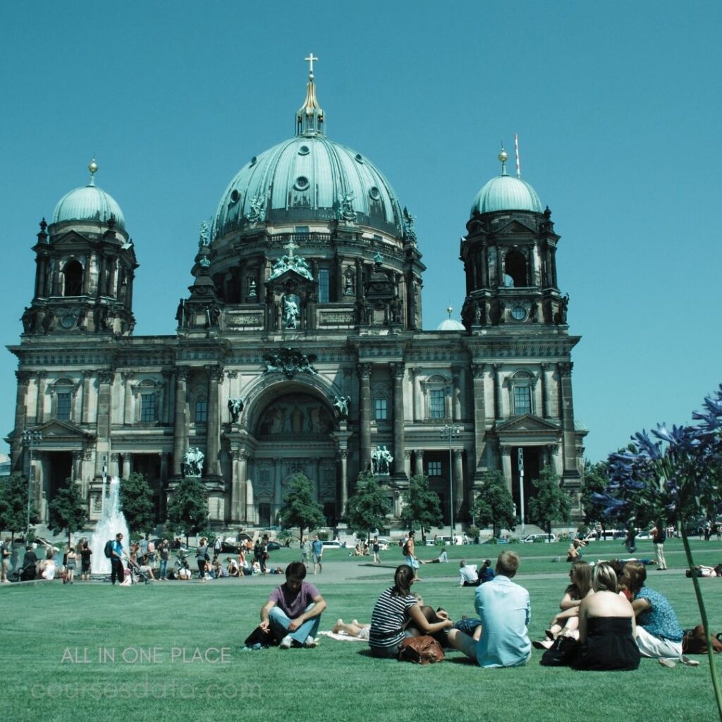 Berlin Cathedral exterior view. Crowd relaxing on the lawn. Clear blue sky above. Green grass and trees surrounding. Dome and towers prominently featured.