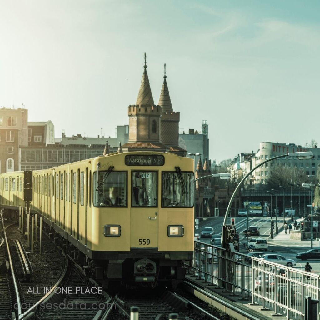 Yellow train at station. Historic tower in background. Urban setting with traffic. Clear blue sky above. Appears to be noon.