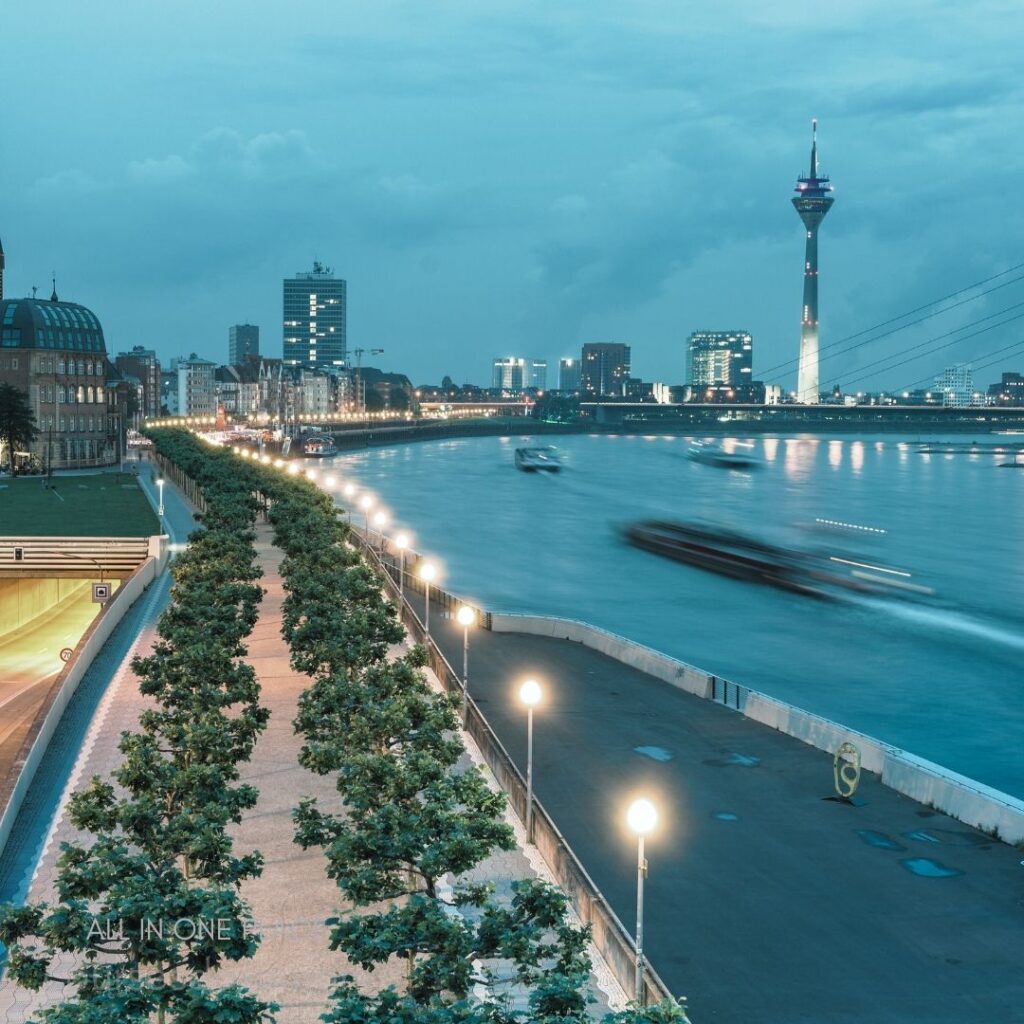 City skyline at twilight. River with blurred boats. Lined trees along the path. TV tower illuminated at night. Buildings reflective in water.