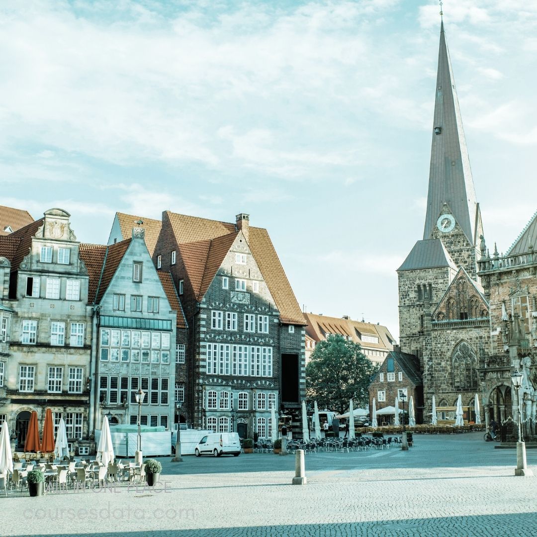 Historic buildings and tower. Paved square with umbrellas. Clear blue sky backdrop. Charming architectural details visible. Quiet atmosphere with few people.