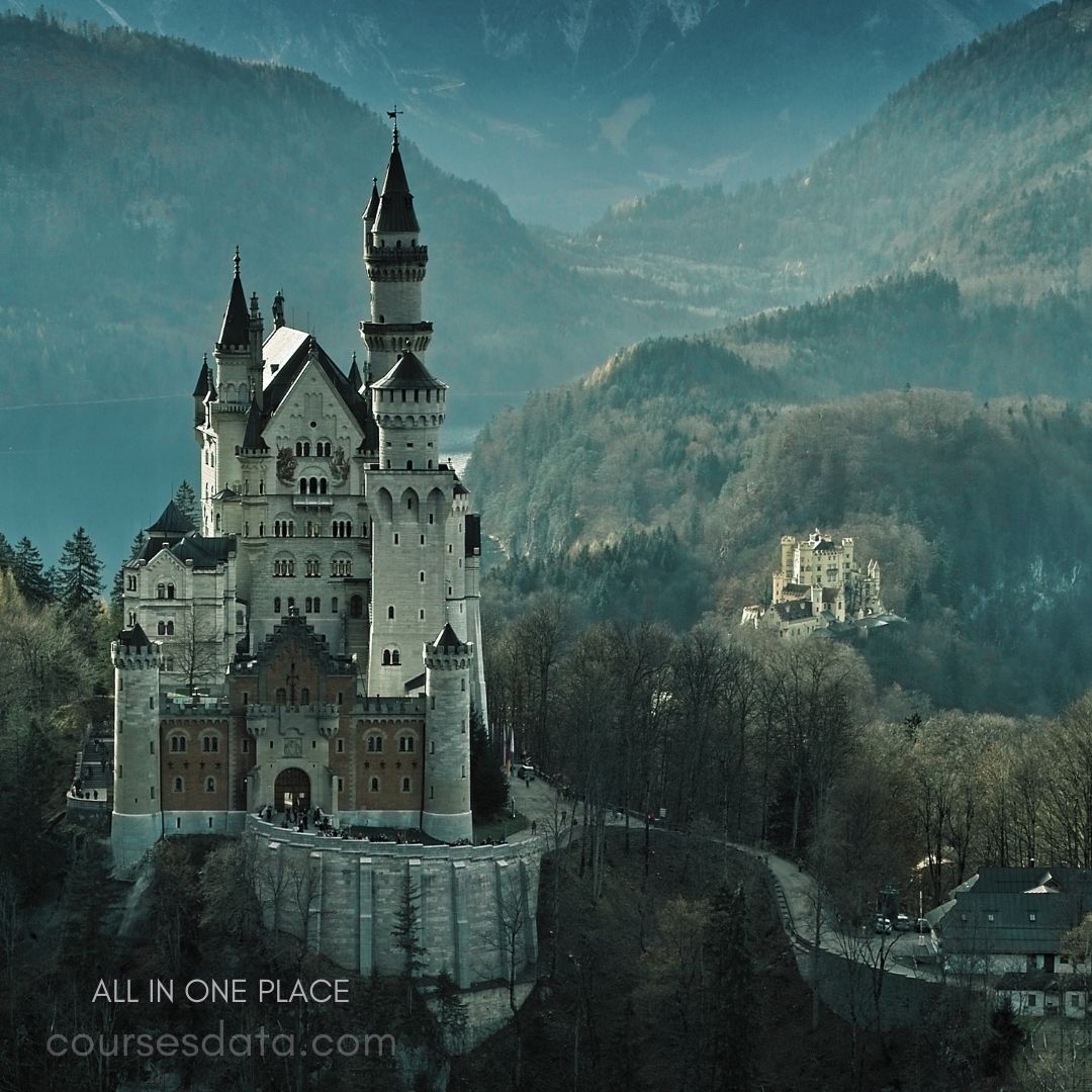 Fairytale castle amidst mountains. Neuschwanstein Castle in focus. Surrounded by dense trees. Distant castle on hilltop. Mist-covered landscape backdrop.