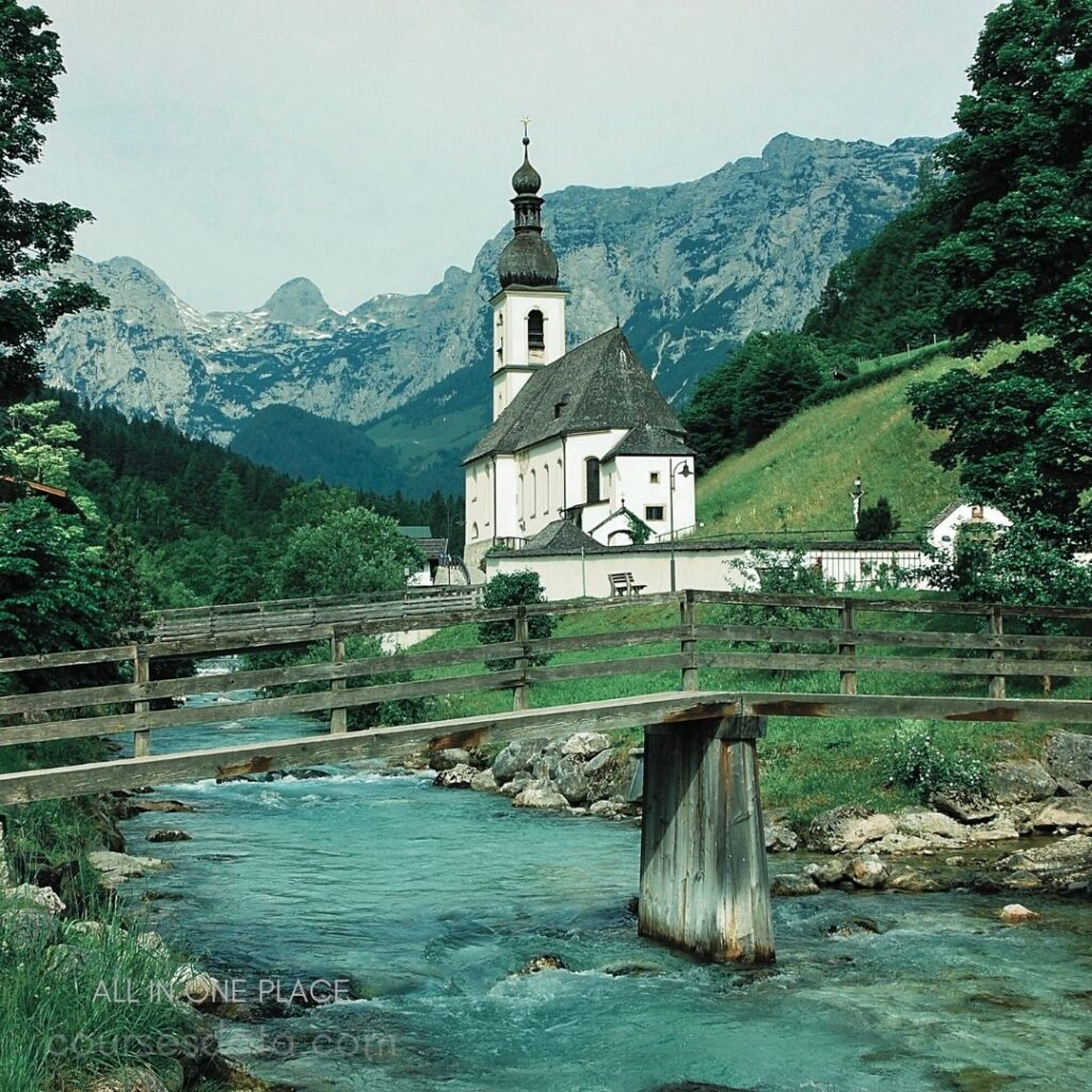 Church beside flowing river. Mountains in background. Wooden bridge crossing water. Lush greenery surrounding area. Peaceful rural landscape scene.