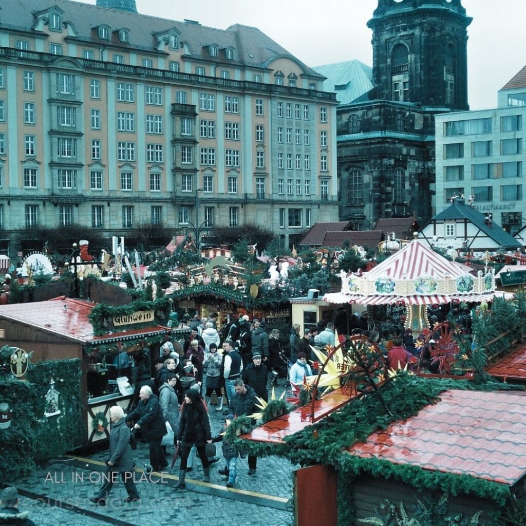 Christmas market bustling with vendors. Colorful stalls decorated with greenery. Historic buildings in the background. Crowd enjoying festive atmosphere. Gray sky over the market.