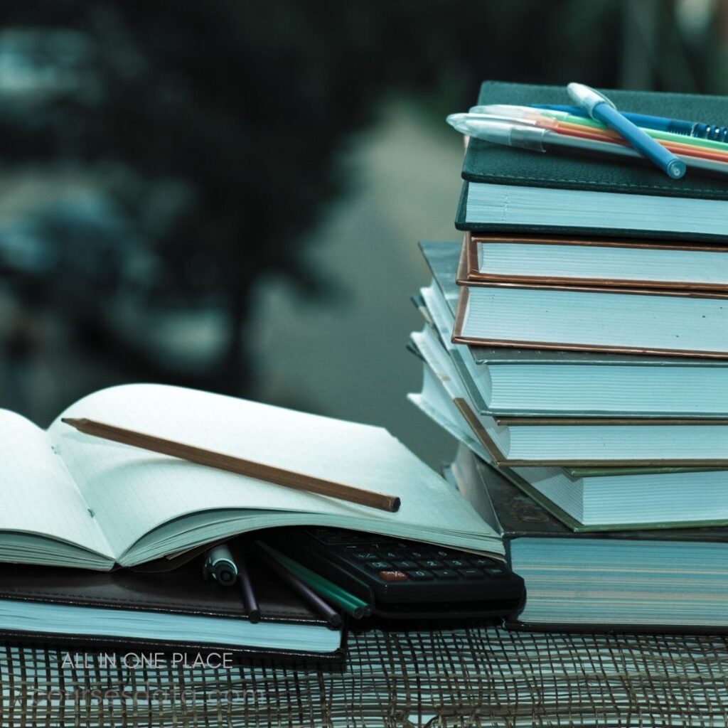 Books stacked on a table. Open notebook with pencil. Calculator and colored pens. Outdoor background blurred softly. Organized study materials displayed.