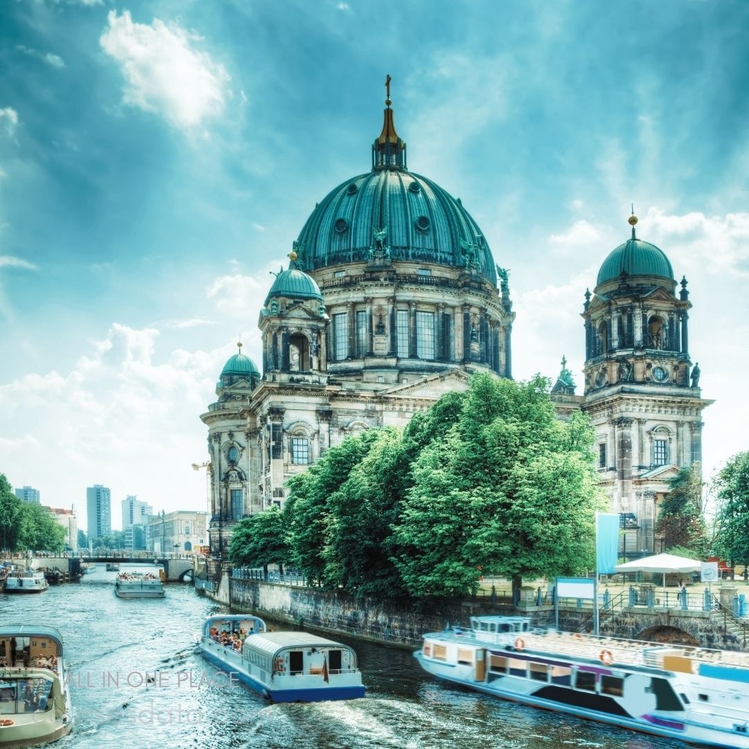 Berlin Cathedral soaring above river. Boats cruising on serene water. Lush trees lining the bank. Bright, cloudy blue sky overhead. Historical architecture with domes.