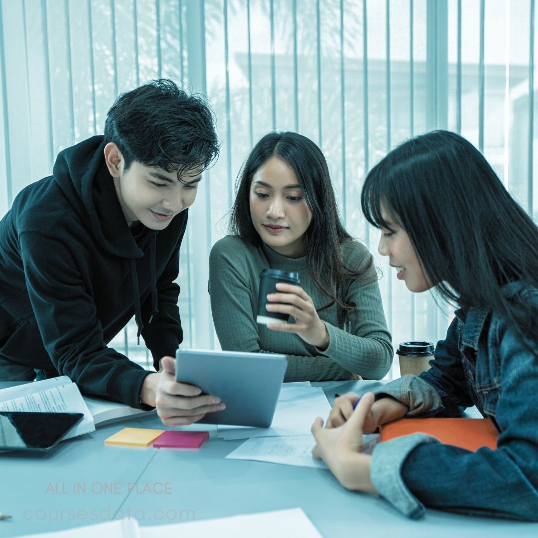 Three students collaborating together. Young man using a tablet. One woman holding a coffee. Colorful sticky notes on table. Bright, modern classroom setting.