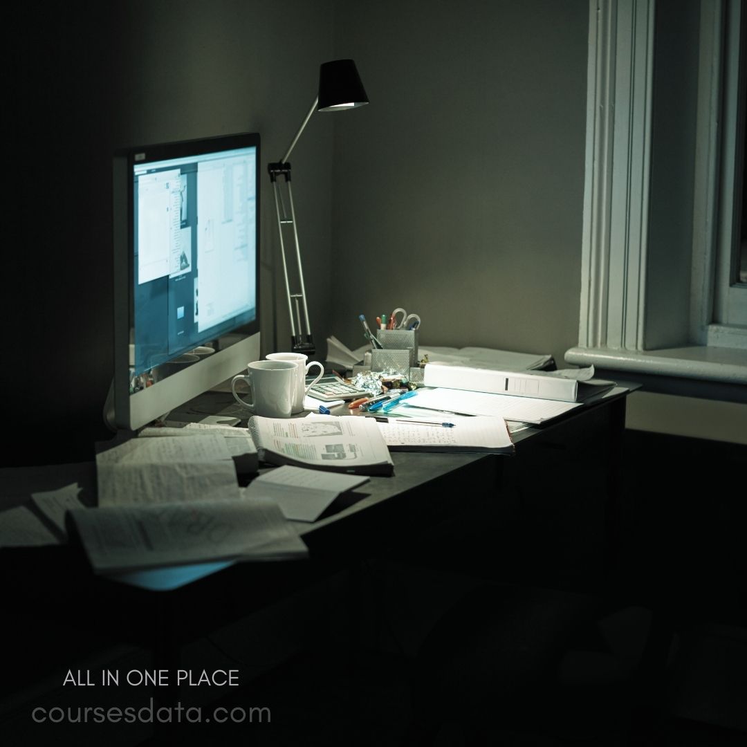 Cluttered desk with monitor. White mug on papers. Desk lamp illuminating workspace. Scattered notes and stationery. Window with dark surroundings.