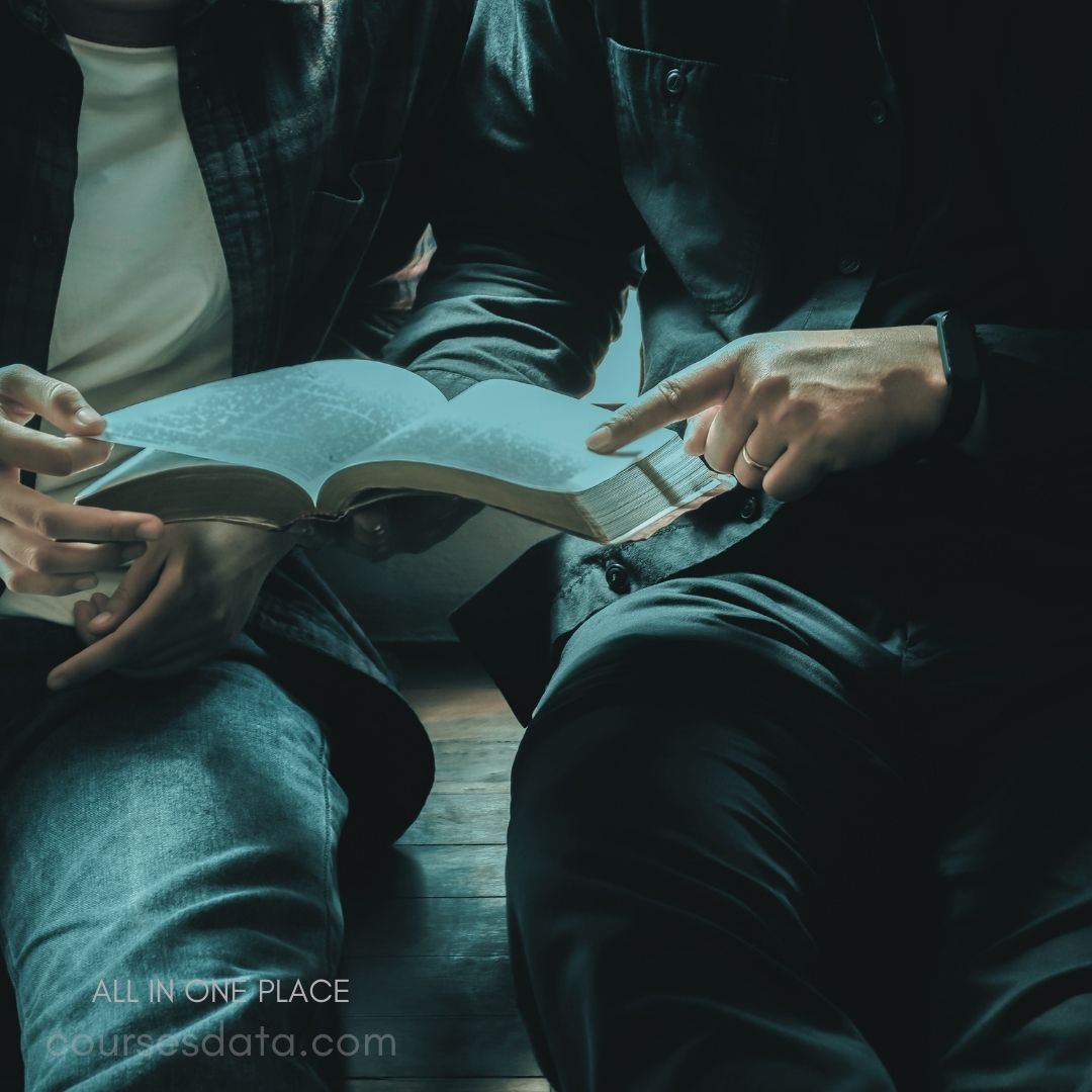 Two hands holding a book. Open pages show text. Cozy seating area with natural light. Dark clothing contrasts with book. Engaged discussion or reading.