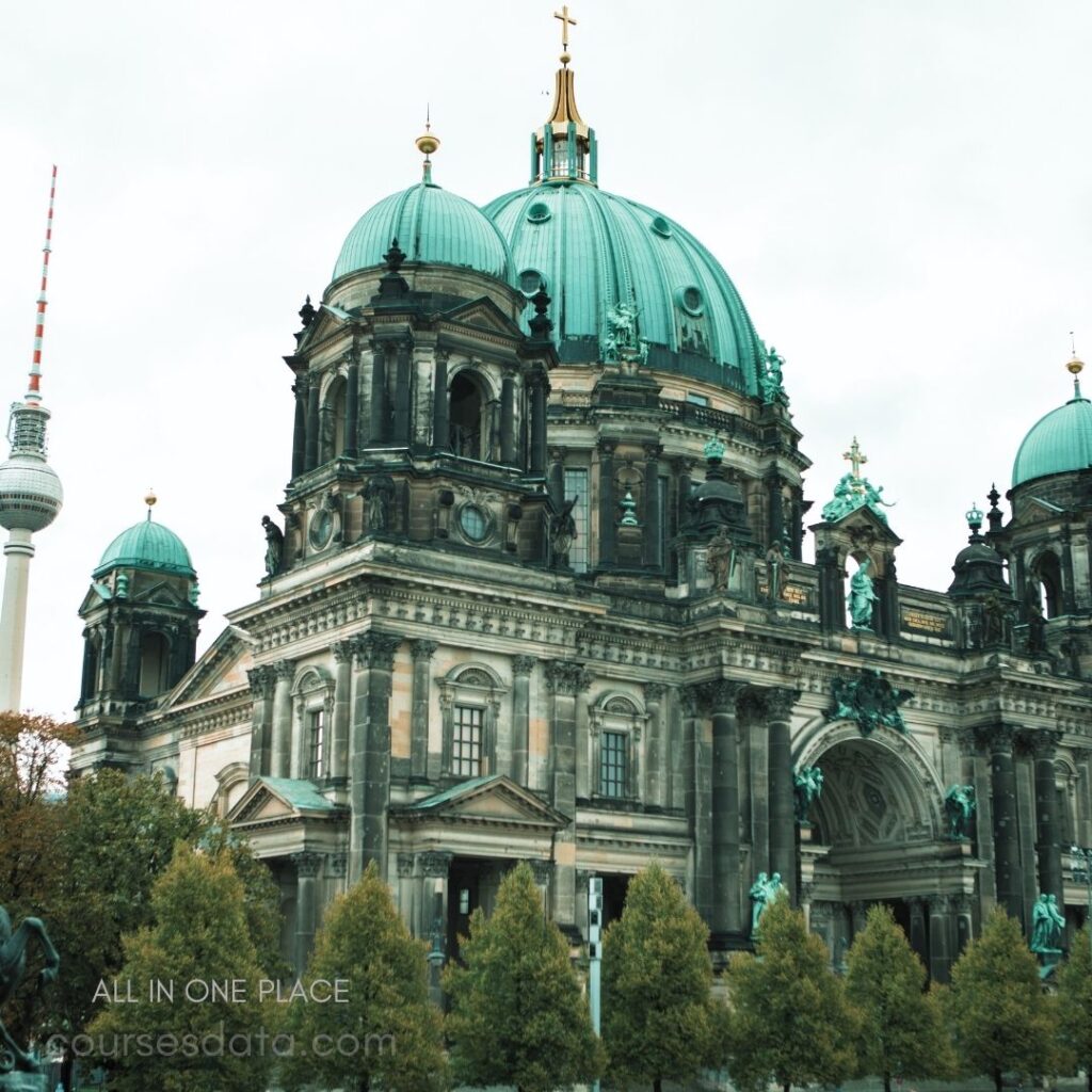 Berlin Cathedral architecture details. Distinctive green copper domes. Television tower in background. Surrounding greenery and trees. Gothic and Baroque features visible.