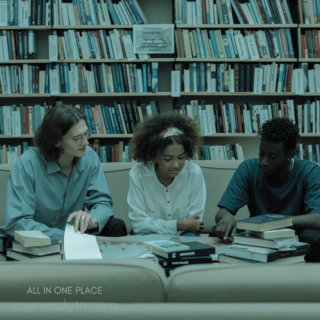 Three students studying together. Books scattered on the table. Library bookshelves in background. Casual, focused expressions displayed. Paperwork laid out between them.