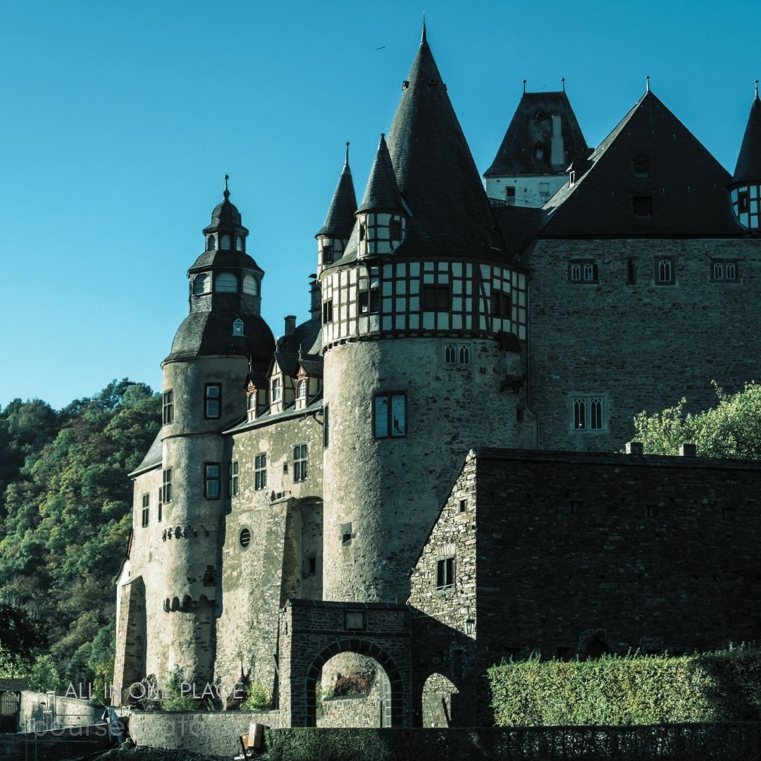 Historic castle with towers. Stone walls and turreted roofs. Surrounded by lush green hills. Clear blue sky backdrop. Vibrant natural scenery.