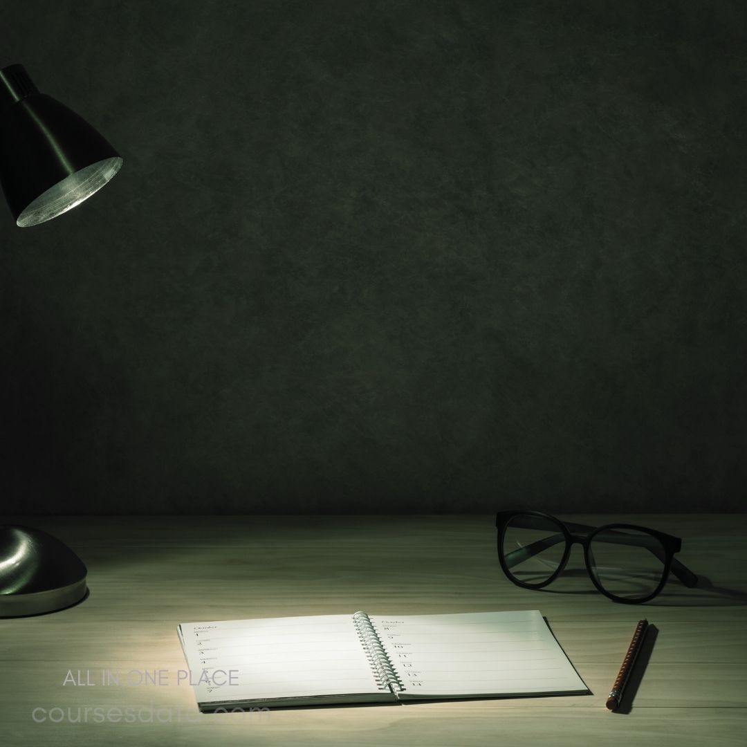 Desk lamp illuminates notebook. Open notebook with blank pages. Black glasses beside the notebook. Wooden table surface underneath items. Dark, moody background ambiance.