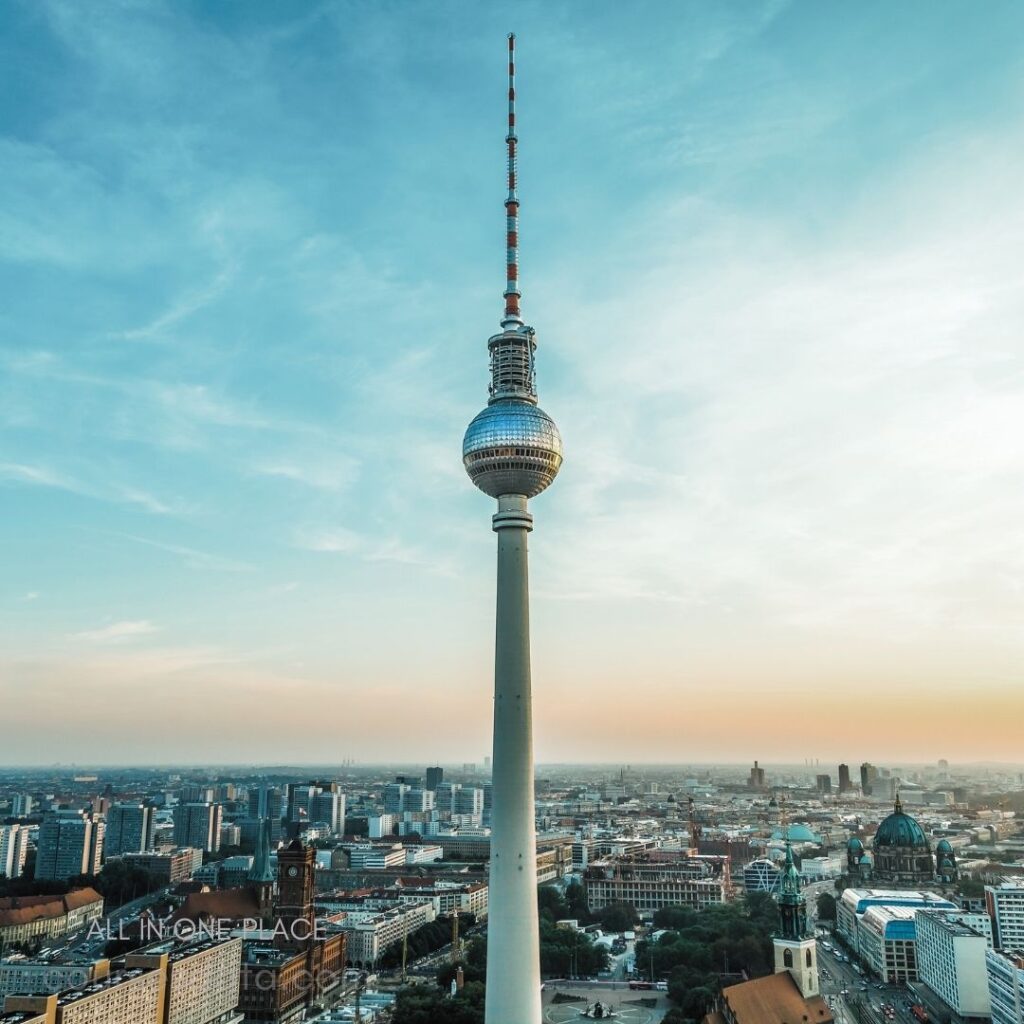 Berlin TV Tower prominently displayed. Blue sky with soft clouds. City skyline in the background. Warm sunset lighting the scene. Historical buildings visible below.