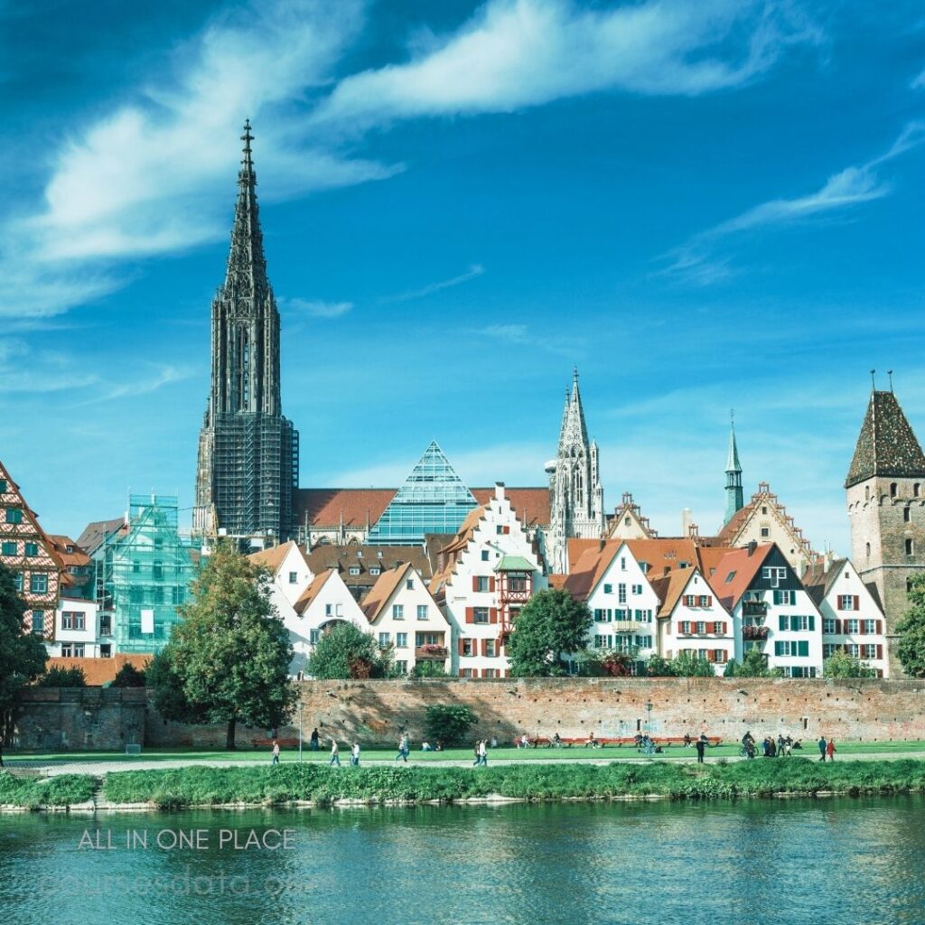 Historic buildings by river. Tall spires dominate skyline. Green trees line waterfront. People walking along path. Clear blue sky overhead.
