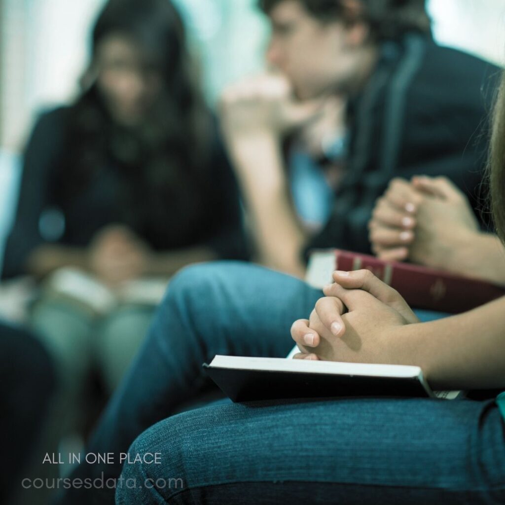 Group praying with books. Hands clasped in prayer. Casual attire, focused expressions. Blurred background, soft lighting. Community gathering, spiritual moment.