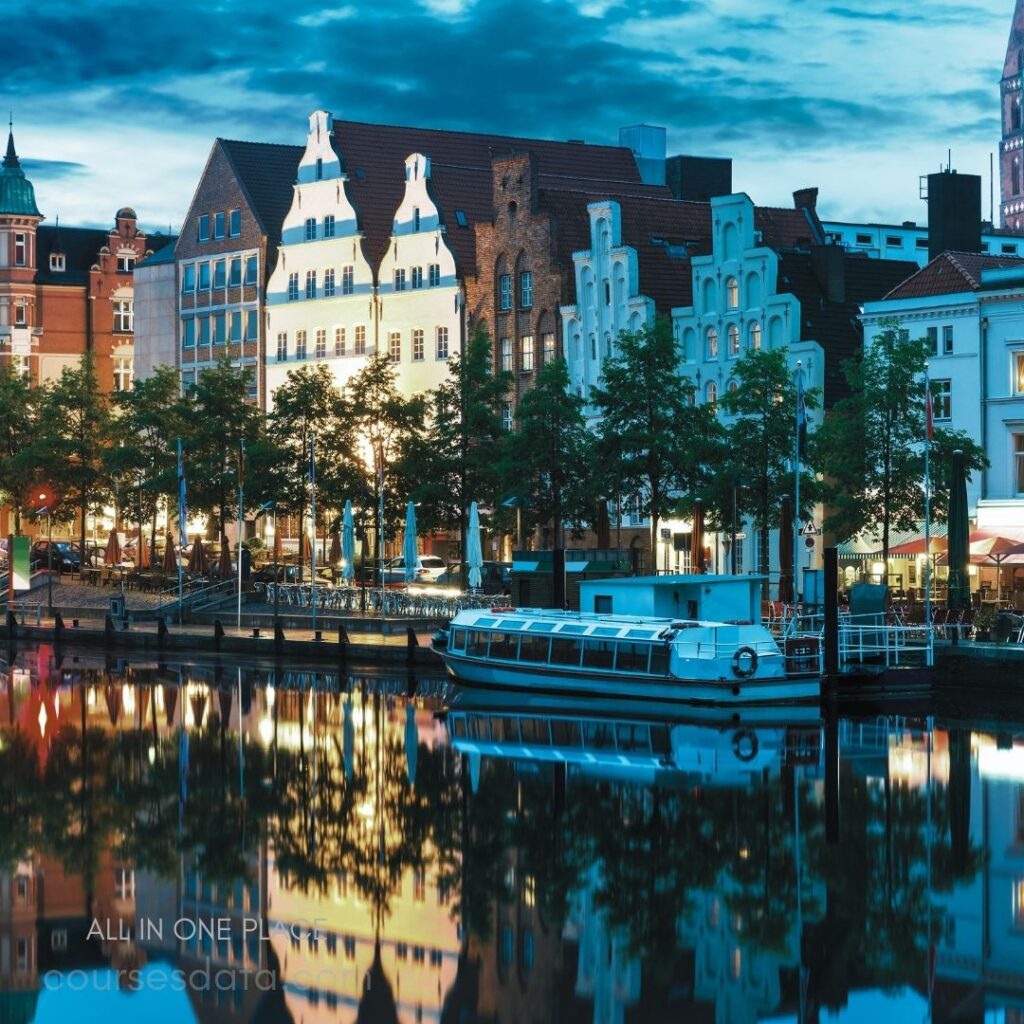 Historic buildings by the water. Reflective calm water surface. Streetlights illuminating evening scene. Boats moored along the dock. Lively atmosphere with outdoor dining.