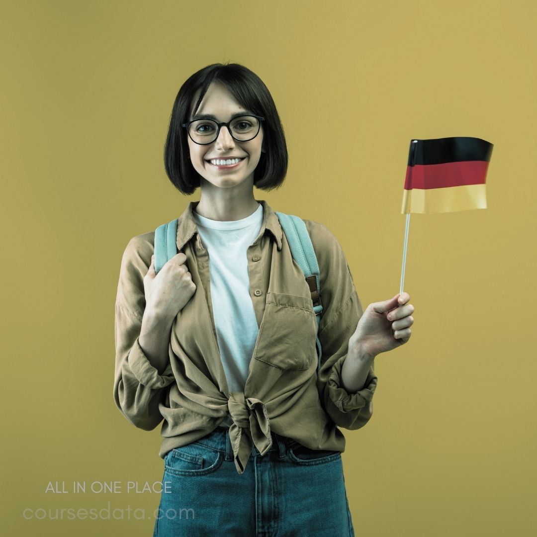 Smiling woman holds German flag. Wearing glasses and casual clothing. Light yellow background. Backpack slung over shoulder. Knot tied in shirt.