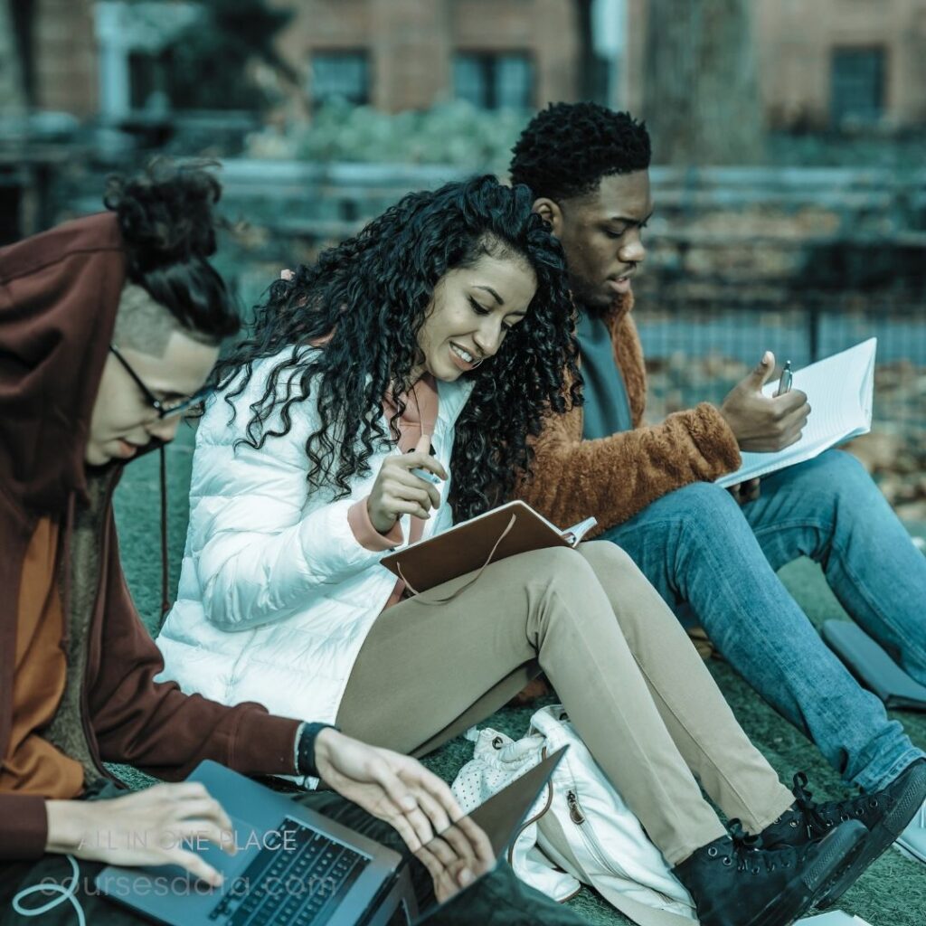 Students studying outdoors together. Woman writing in notebook. Man using laptop. Man taking notes, focused. Autumn leaves scattered around.