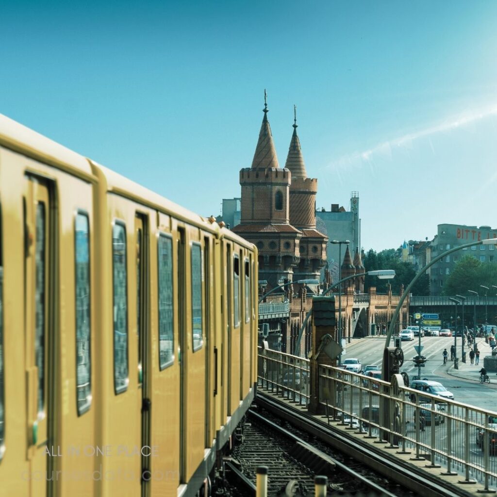 Yellow train on elevated tracks. Historic tower in background. Clear blue sky overhead. Urban scene with pedestrians. Buildings lining the streets.